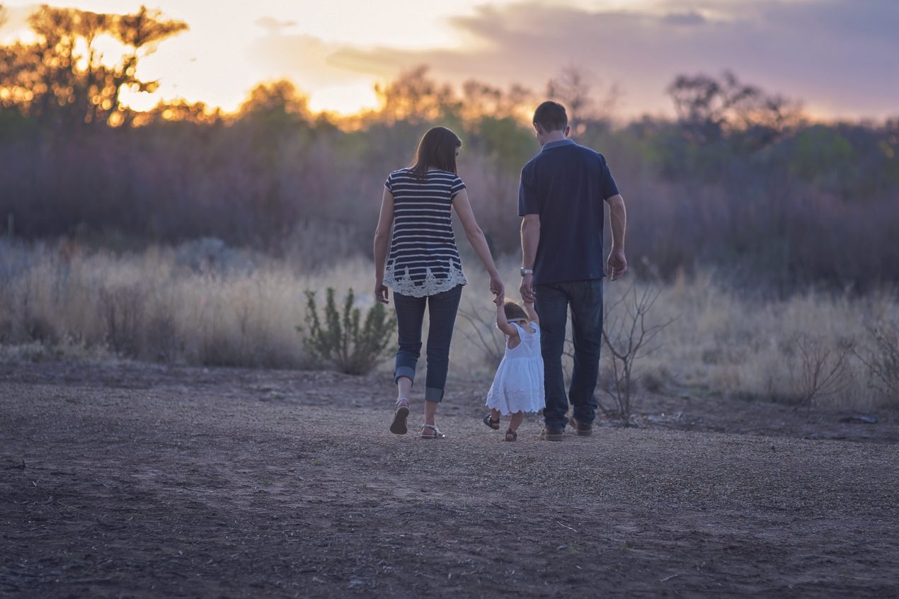 This image shows a family which includes a young mother, father, and toddler in a white dress, walking into the desert against a setting sun, facing the sunset. The mother, father, and child hold hands as a they walk away, with the child in between the two adults.